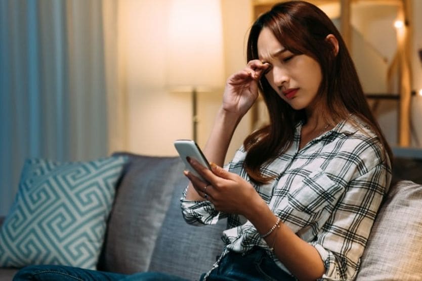 Young woman with eye pain looking at smartphone