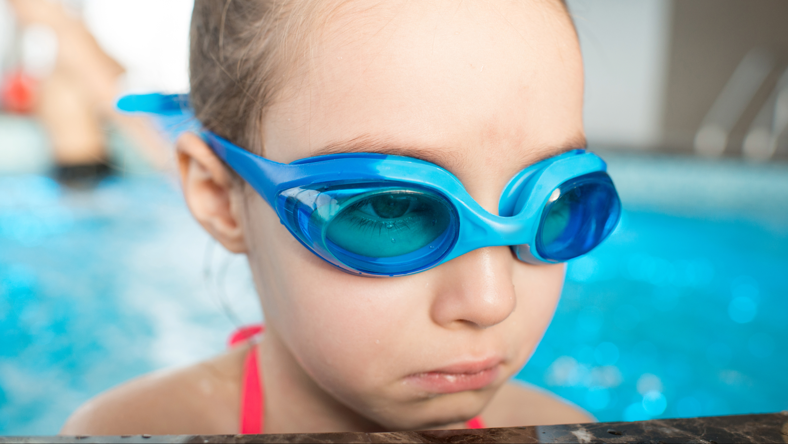 Little girl wearing swim goggles looks sad at the side of a pool