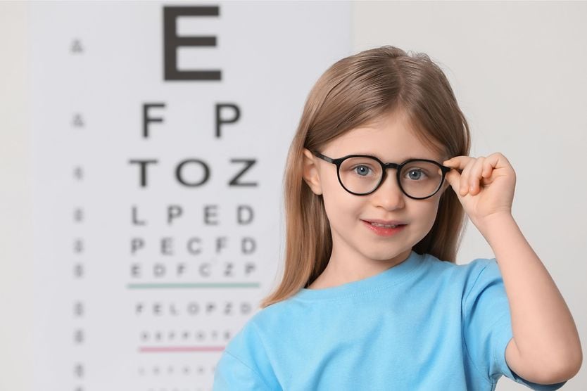 Young girl wearing glasses for myopia standing in front of a Snellen eye chart.