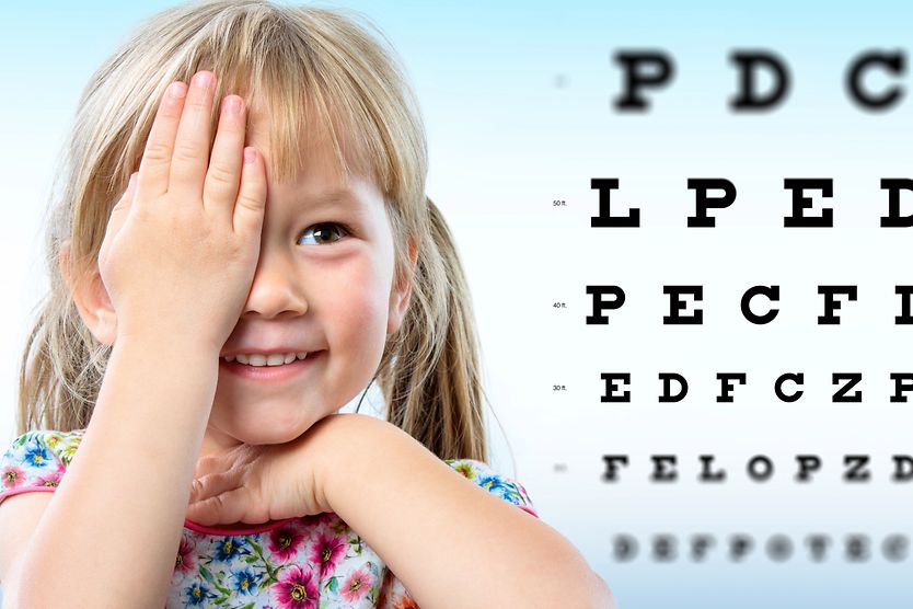 A young girl with myopia covering one eye with her hand in front of a Snellen eye chart.