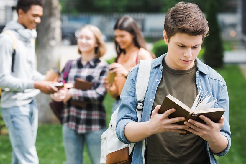 Selective focus of teenager reading book with friends on background.