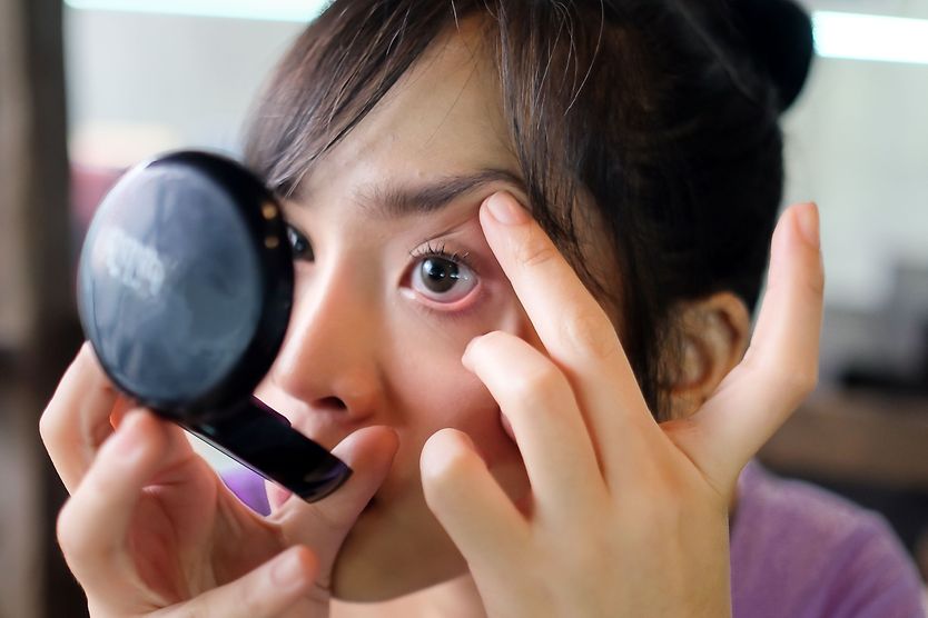 How to safely remove something from your eye woman looking at a compact mirror trying to get something out of her eye