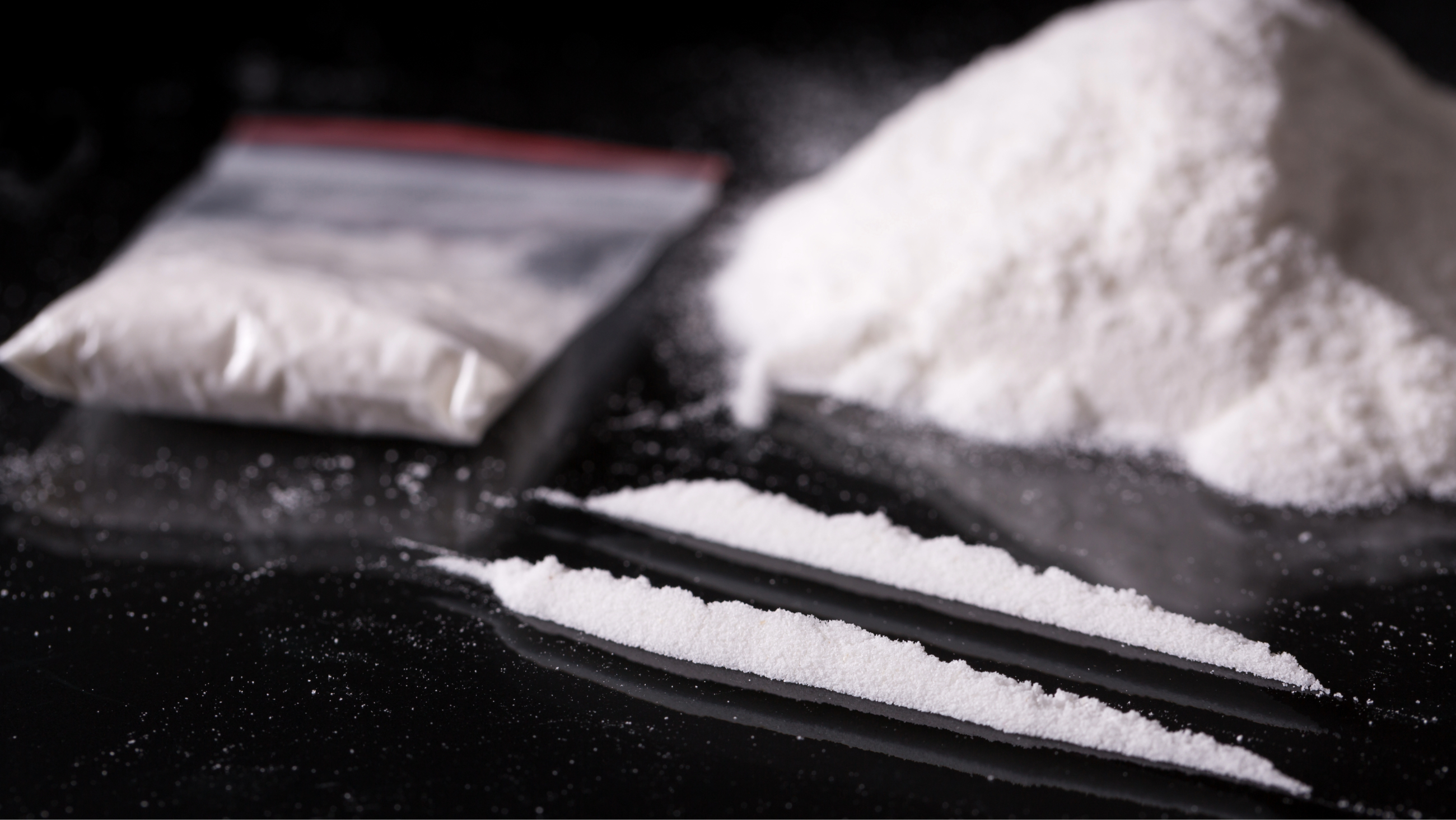 Plastic packet, two lines and pile of cocaine on black background, closeup.