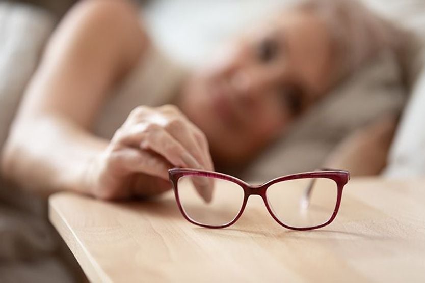 Does age affect eyesight after age 40? Mature woman lying in bed in the background while holding onto her eyeglasses on the nightstand