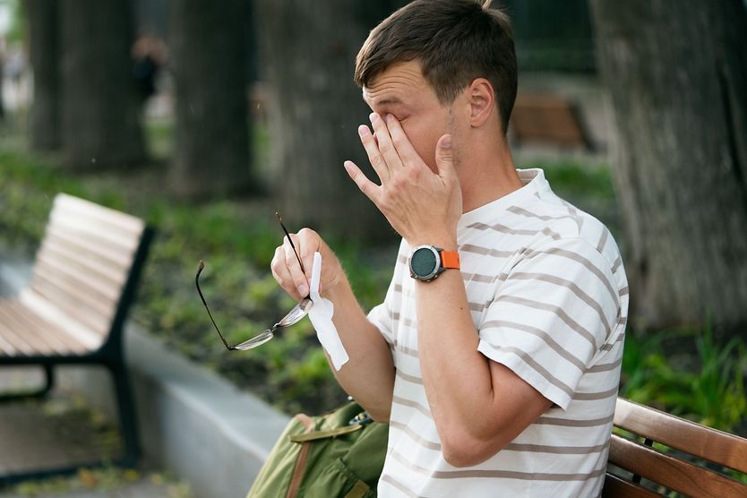 A man outside in nature holding his glasses and a tissue in his hand as he rubs his eye due to allergies.