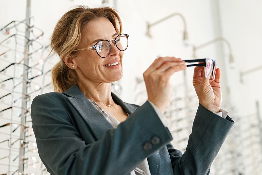 12 glasses that make you look younger Woman chooses between two glasses in an optician's shop.