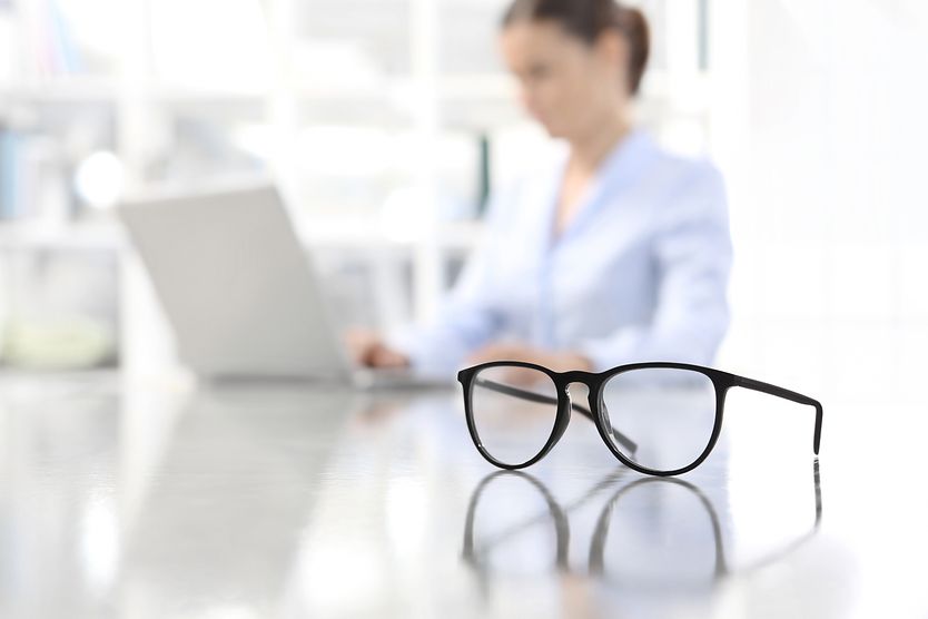 A pair of monovision glasses sitting on a desk next to a woman working at her computer. A pair of monovision glasses sitting on a desk next to a woman working at her computer.