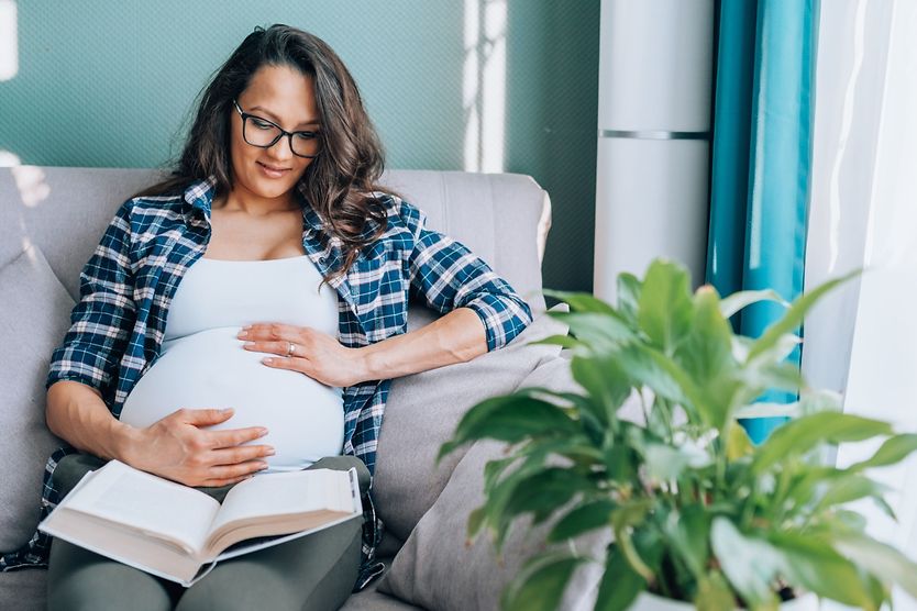 Lasik when pregnant. Relaxed dark-haired pregnant woman in glasses and home wear sit on comfortable grey couch with pillows in living room, touching her tummy, smiling and relaxing while reading book at home.