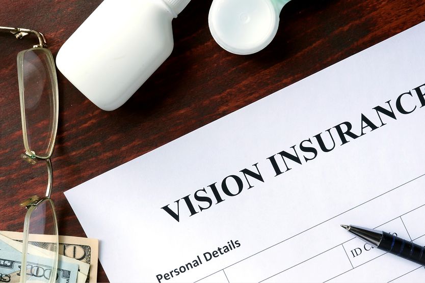 Eyeglasses sitting on a desk next a pen and paperwork labeled as "Vision Insurance." Eyeglasses sitting on a desk next a pen and paperwork labeled as "Vision Insurance."