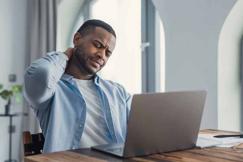Uomo che usa un portatile si massaggia il collo per alleviare il fastidio, evidenziando una scarsa ergonomia della postazione e l'affaticamento visivo dovuto allo schermo.