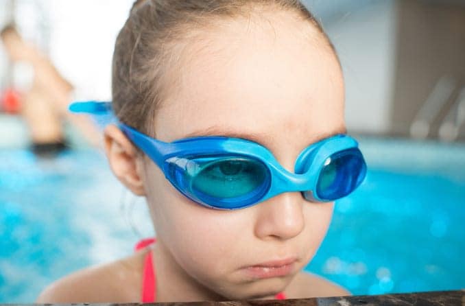 Little girl wearing swim goggles looks sad at the side of a pool