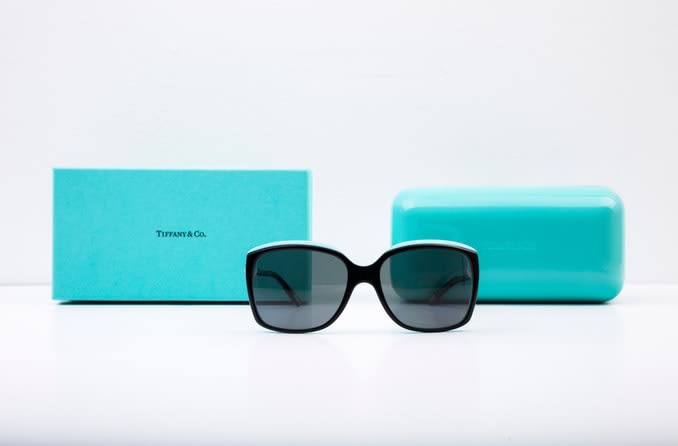 A pair of black Tiffany sunglasses are displayed in front of the iconic turquoise blue Tiffany box and sunglasses case.