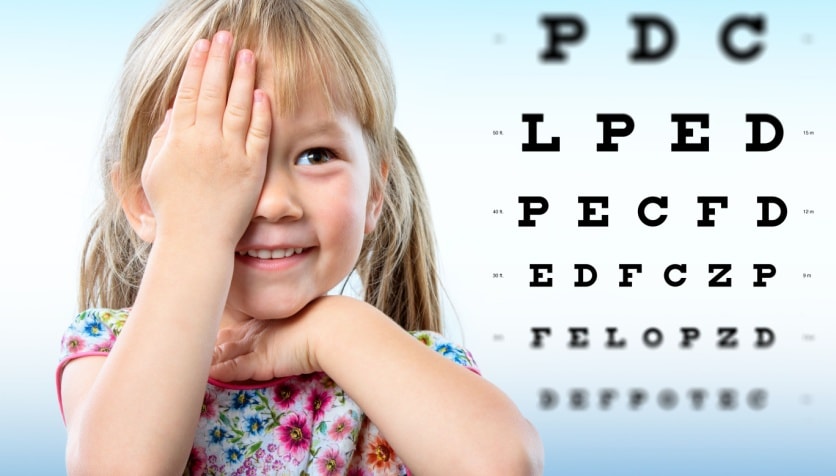 A young girl with myopia covering one eye with her hand in front of a Snellen eye chart.