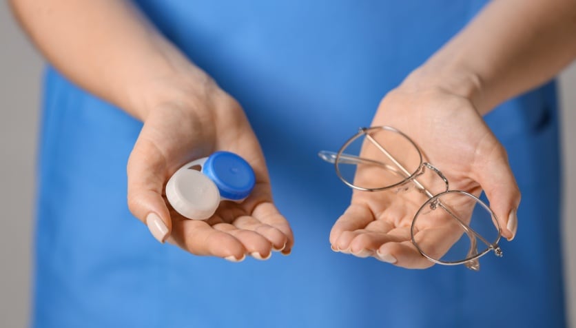 An optician holding contact lenses in a contact lens case in one hand and glasses in her other hand.