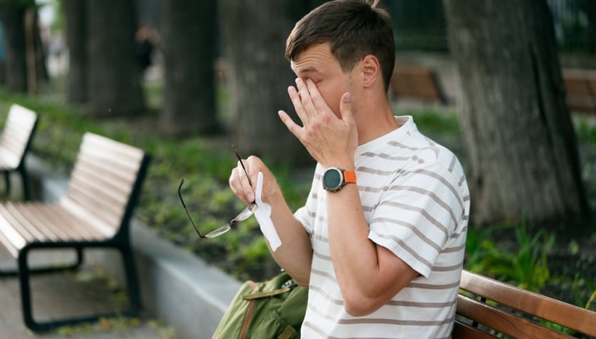 A man outside in nature holding his glasses and a tissue in his hand as he rubs his eye due to allergies.