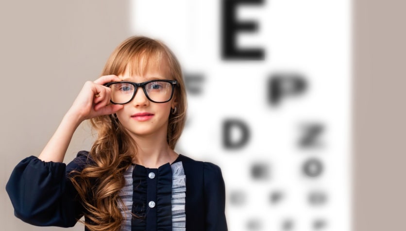 Young girl wearing glasses for myopia standing in front of a Snellen eye chart.