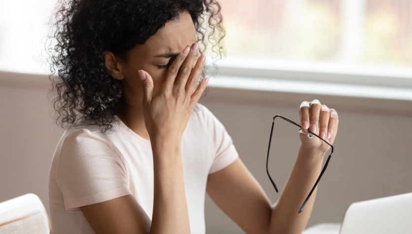 Woman sitting in front of a computer holding her glasses with her hand to her head due to headache.