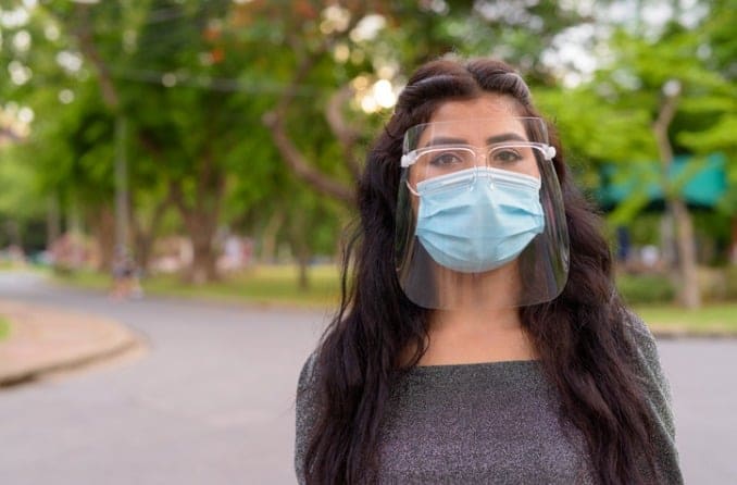Young woman with long brown hair is wearing both a clear face shield and a face mask.