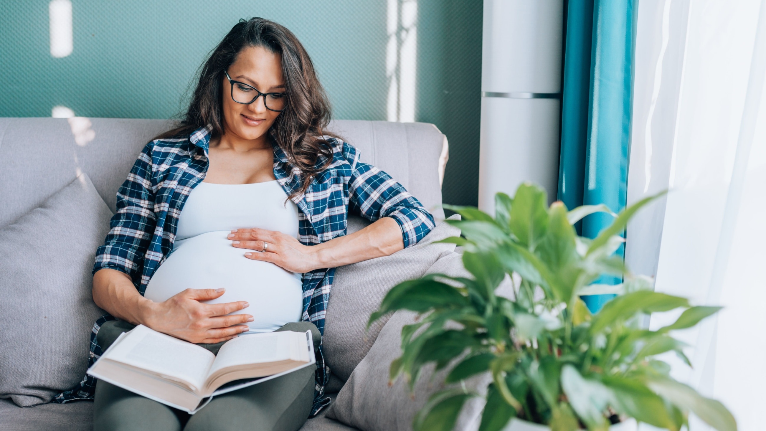 Relaxed dark-haired pregnant woman in glasses and home wear sit on comfortable grey couch with pillows in living room, touching her tummy, smiling and relaxing while reading book at home.