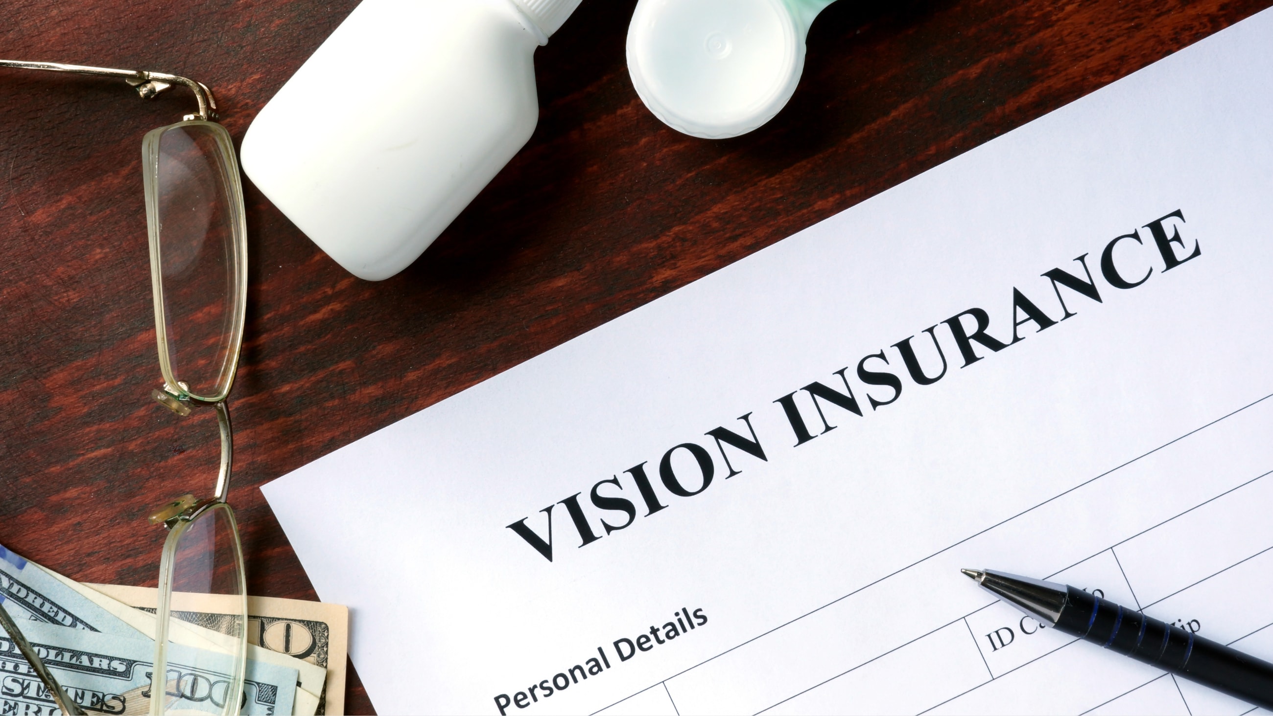 Eyeglasses sitting on a desk next a pen and paperwork labeled as "Vision Insurance." Eyeglasses sitting on a desk next a pen and paperwork labeled as "Vision Insurance."