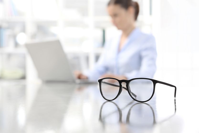 A pair of monovision glasses sitting on a desk next to a woman working at her computer. A pair of monovision glasses sitting on a desk next to a woman working at her computer.