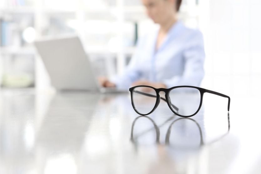 A pair of monovision glasses sitting on a desk next to a woman working at her computer. A pair of monovision glasses sitting on a desk next to a woman working at her computer.