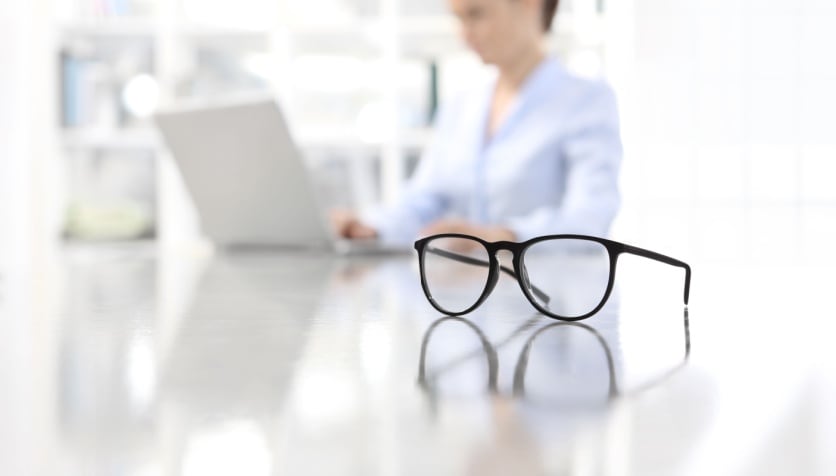 A pair of monovision glasses sitting on a desk next to a woman working at her computer.