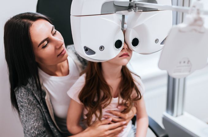 Ragazza seduta sulle ginocchia della madre per eseguire un test oculistico e definire la sua acutezza visiva Ragazza seduta sulle ginocchia della madre per eseguire un test oculistico e definire la sua acutezza visiva