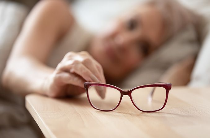Mature woman lying in bed in the background while holding onto her eyeglasses on the nightstand