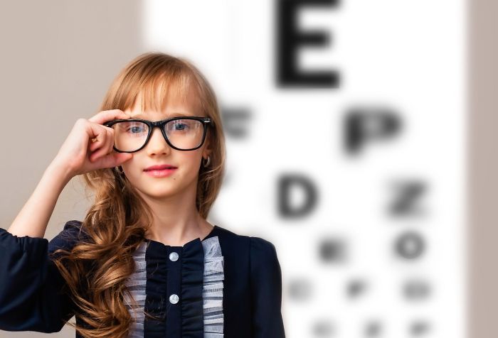 Young girl wearing glasses for myopia standing in front of a Snellen eye chart.