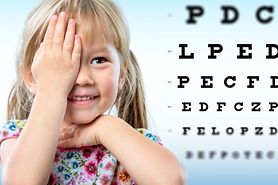 A young girl with myopia covering one eye with her hand in front of a Snellen eye chart.