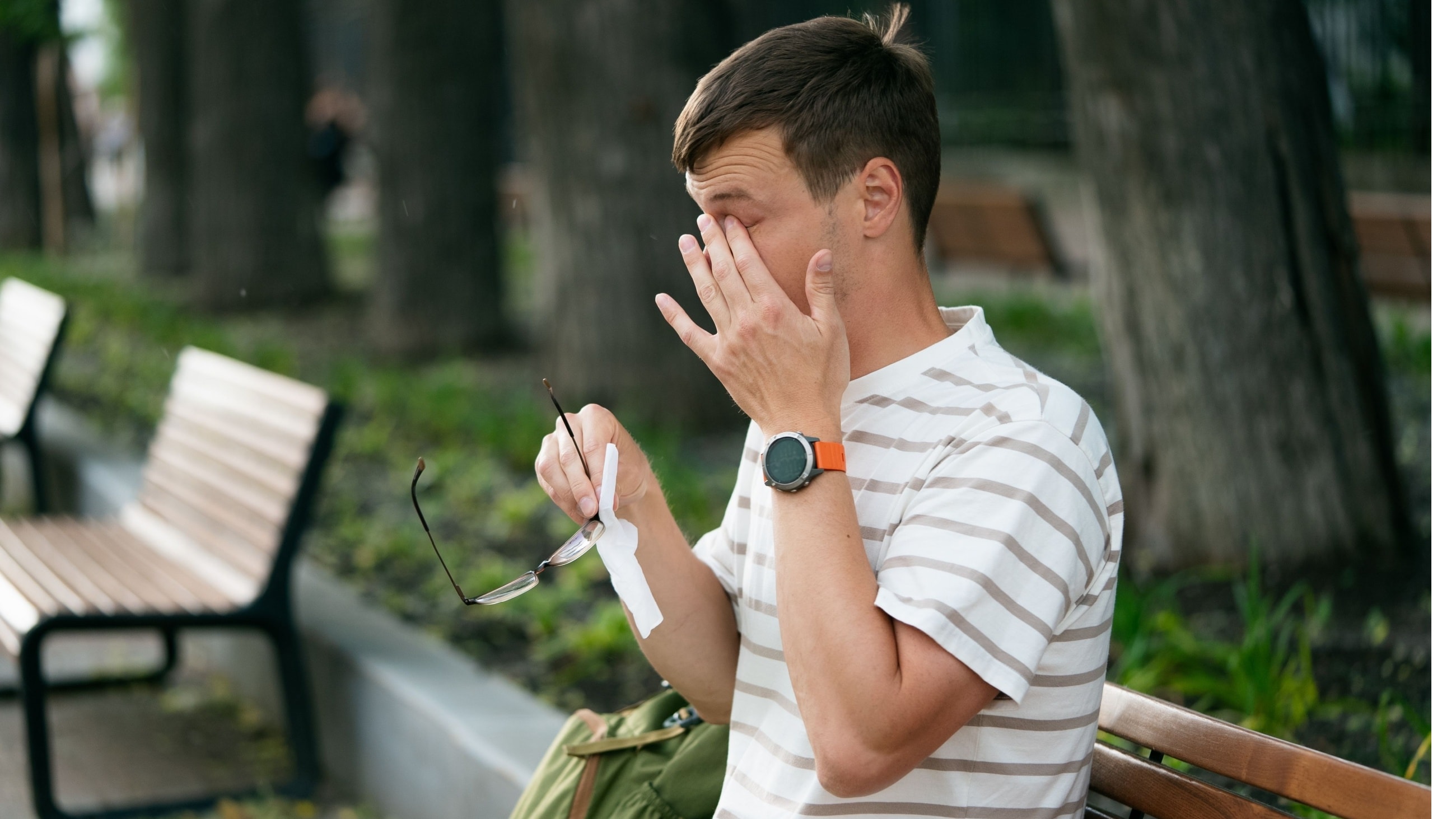 man rubbing eyes with allergies sitting on a park bench