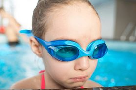 Little girl wearing swim goggles looks sad at the side of a pool