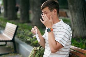 A man outside in nature holding his glasses and a tissue in his hand as he rubs his eye due to allergies.